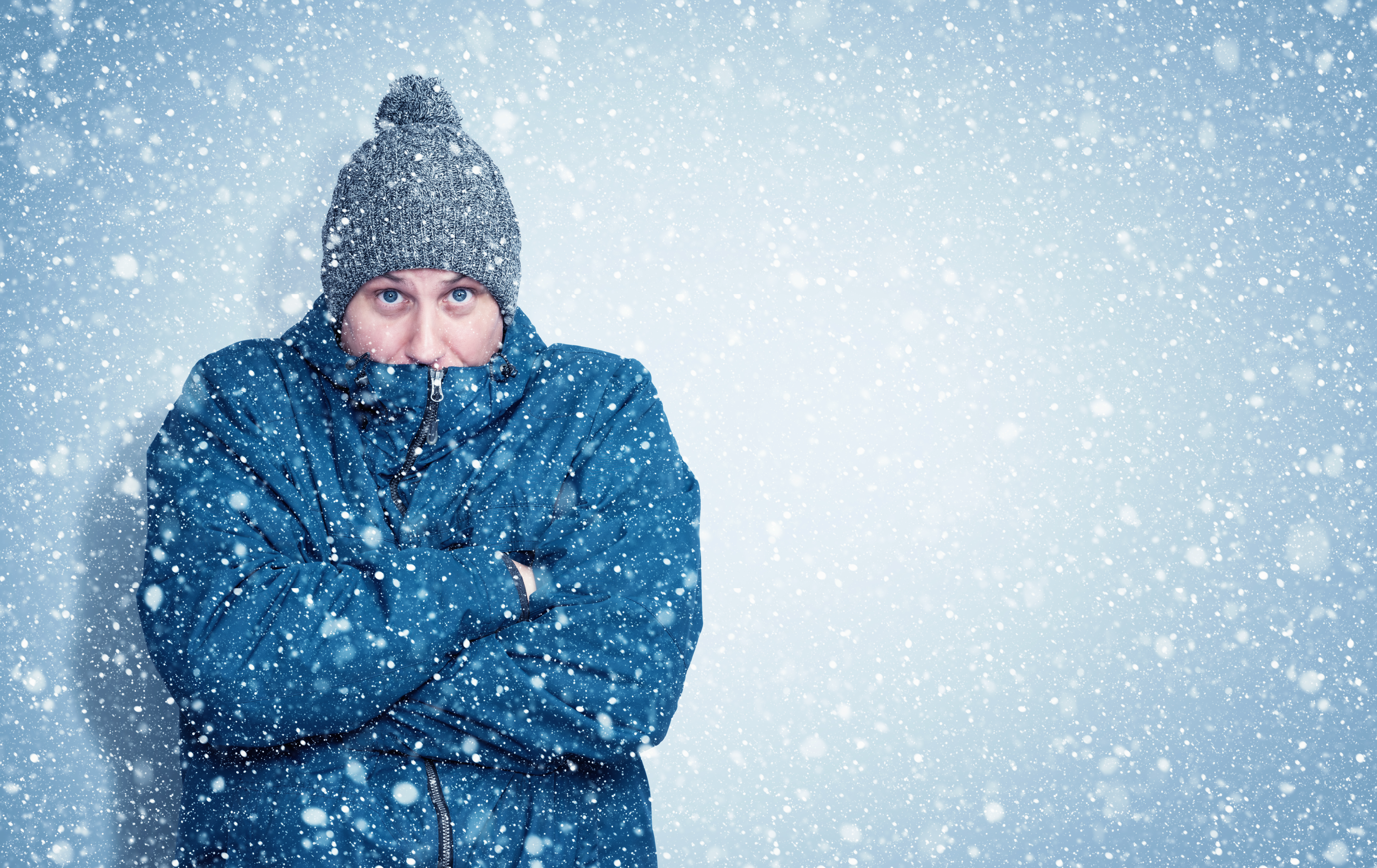 Frozen man in a blue jacket and hat stands against the wall, sno