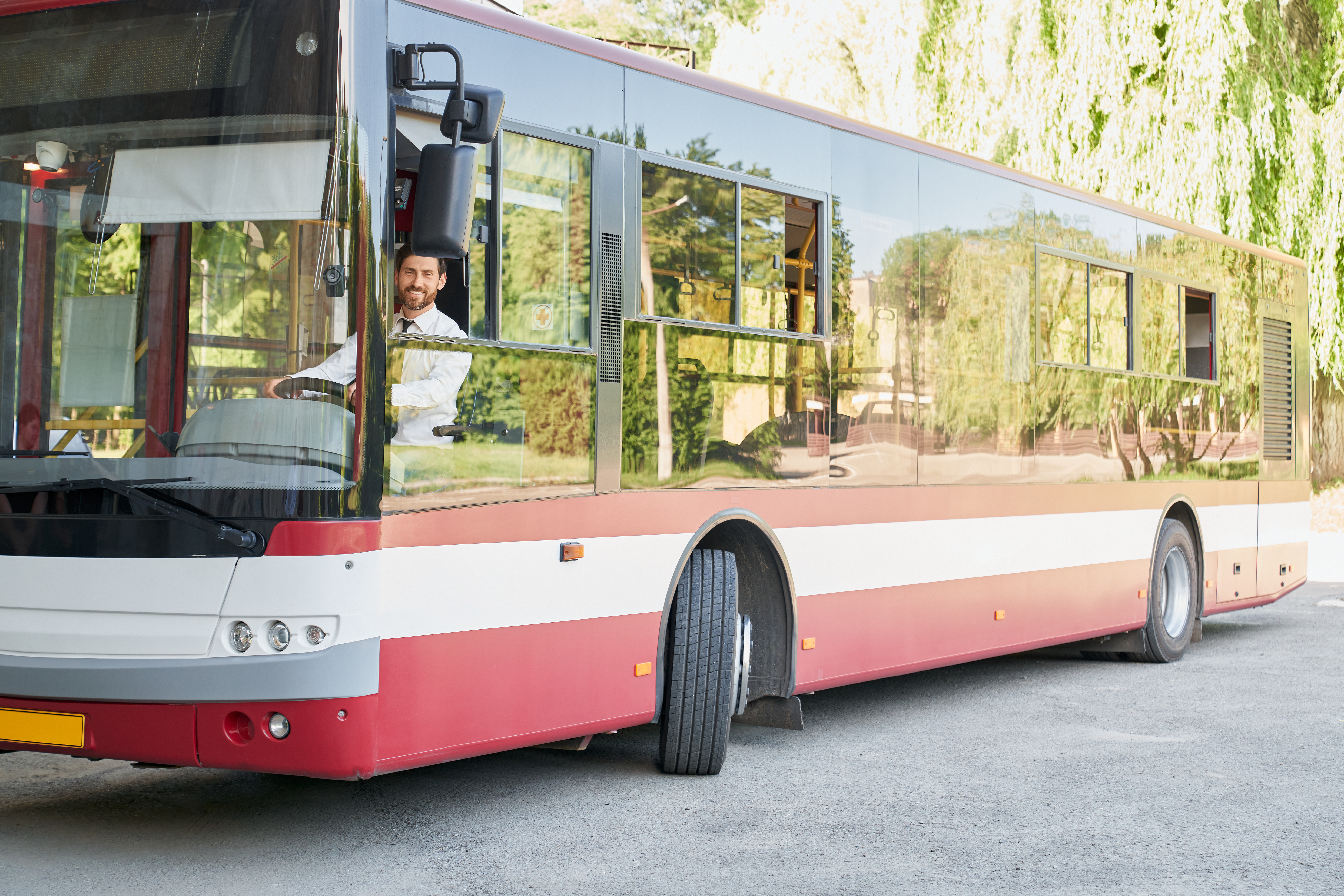 Elegant driver sitting in shuttle bus, smiling to camera at summer day. Front view of happy man sitting on driver's seat, working, going on route way in city. Concept of transportation, work, traffic.