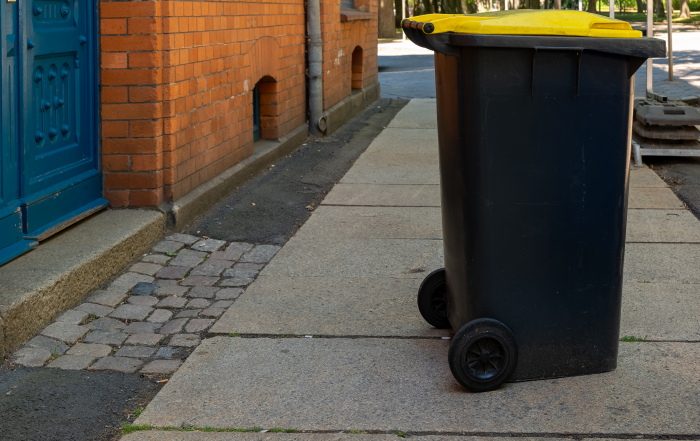 A black trash bin with a bright yellow lid sits on a smooth sidewalk beside a red brick building. Trees provide shade in the background, indicating a calm, sunny day.